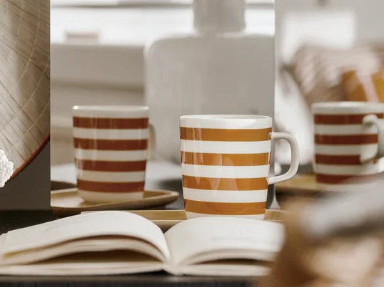 Der Oiva Tasaraita Becher mit Henkel von Marimekko in der Ambienteansicht. Das Wollweiss und das Braun gehen zusammen ein harmonisches Streifenmuster ein.