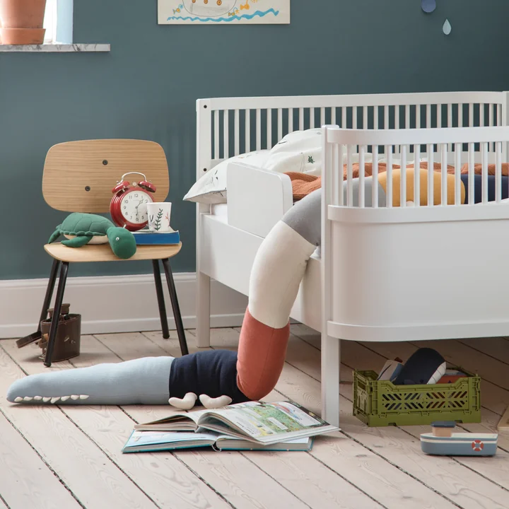 Das Sleepy Croc Kissen und der Oakee Stuhl von Sebra neben dem weissen Kinderbett