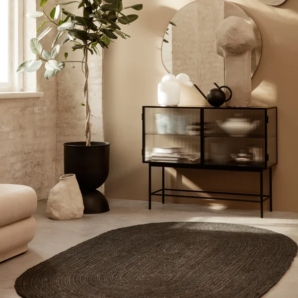Der Eternal Oval Jute Teppich von Ferm Living vor einem Sideboard und neben einer hohen Zimmerpflanze