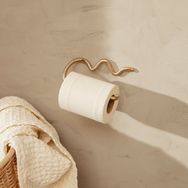 Der Curvature Toilettenpapierhalter von ferm Living an einer hellen Steinwand neben einem Rattan-Korb